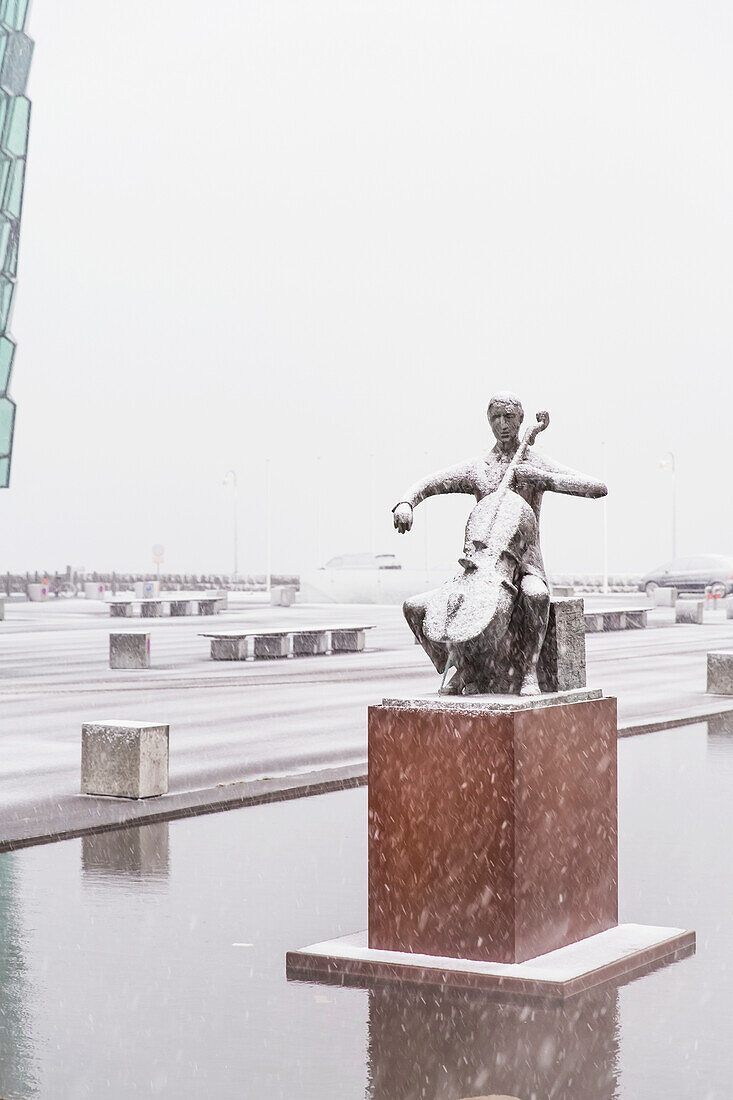 'A statue of cellist Erling Blondal Bengtsson by sculptor Olof Palsdottir sits outside the Harpa Public Concert Hall, snow falling and covering the statue; Reykjavik, Iceland'