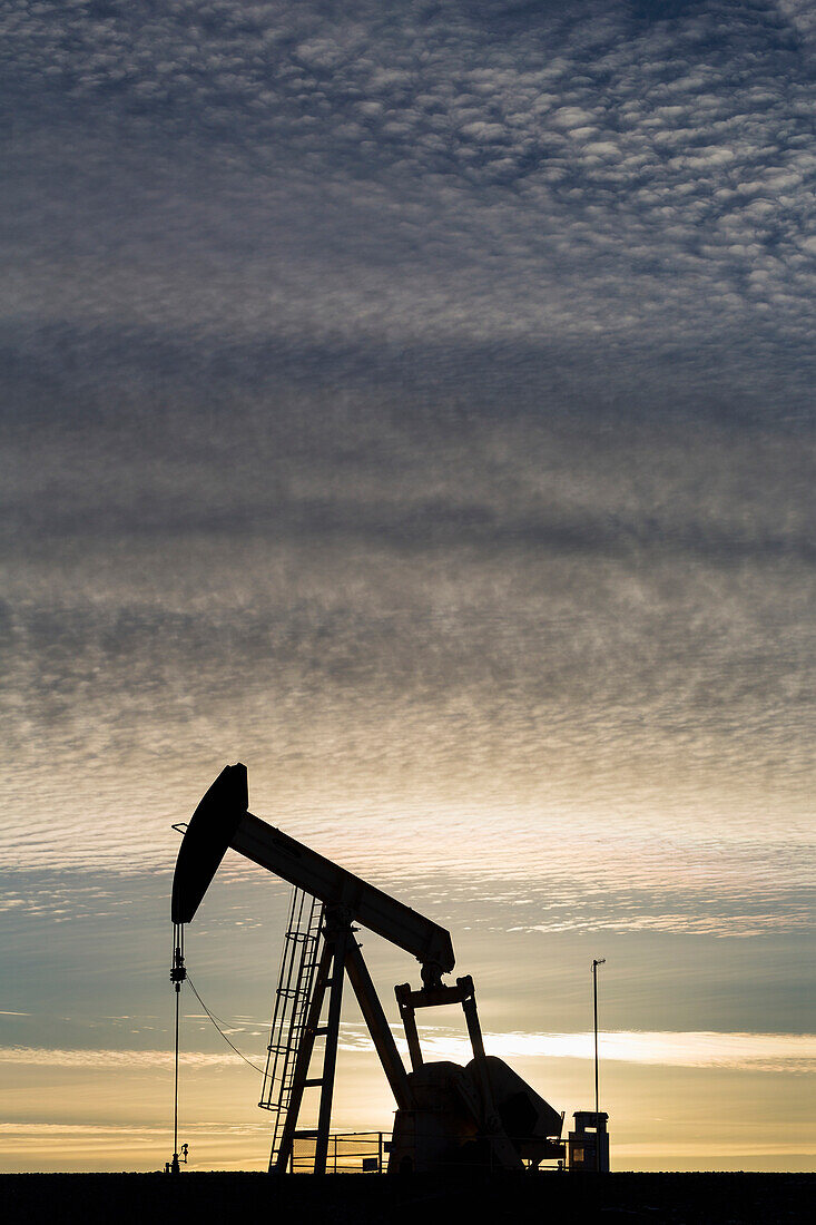 'Silhouette of a pump jack at sunrise with the sun glowing on the horizon and colourful clouds in the sky; Alberta, Canada'