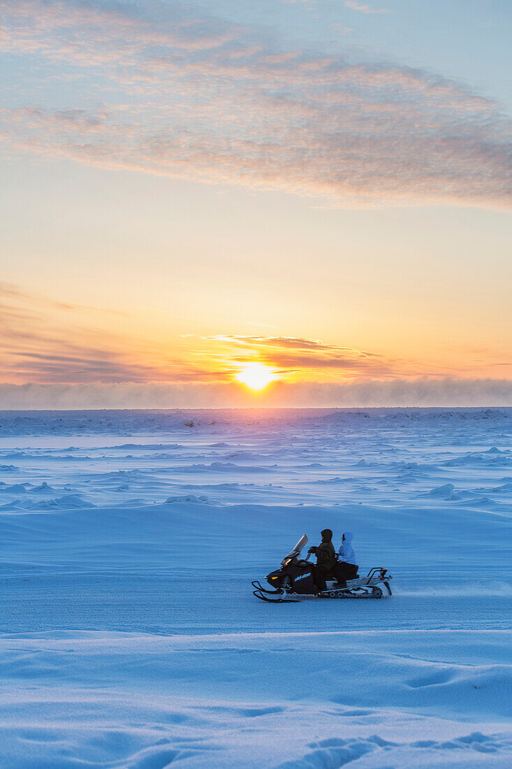 Two native Alaskans drive a snow mobile through a stark snow covered landscape at sunset, Barrow, North Slope, Arctic Alaska, Winter