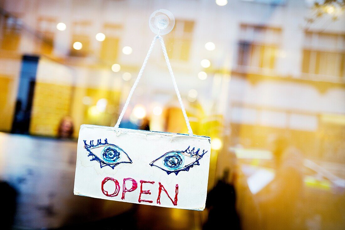 A sign hanging on the glass door of a business, with reflections of the buildings, OPEN with the text and drawn two eyes, London, England, UK, Europe.