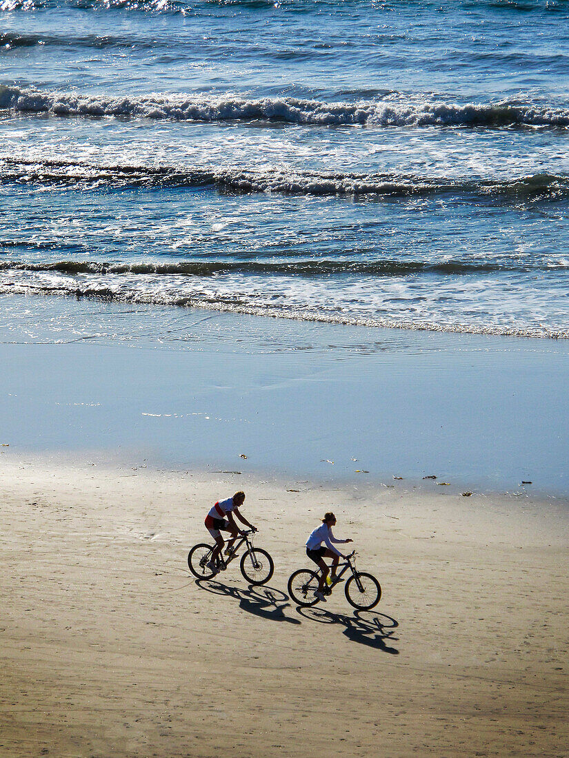 Early morning sunlight throws the shadows of two bicycle riders at Crystal Cove in Newport Beach, CA. Note heavy surf.