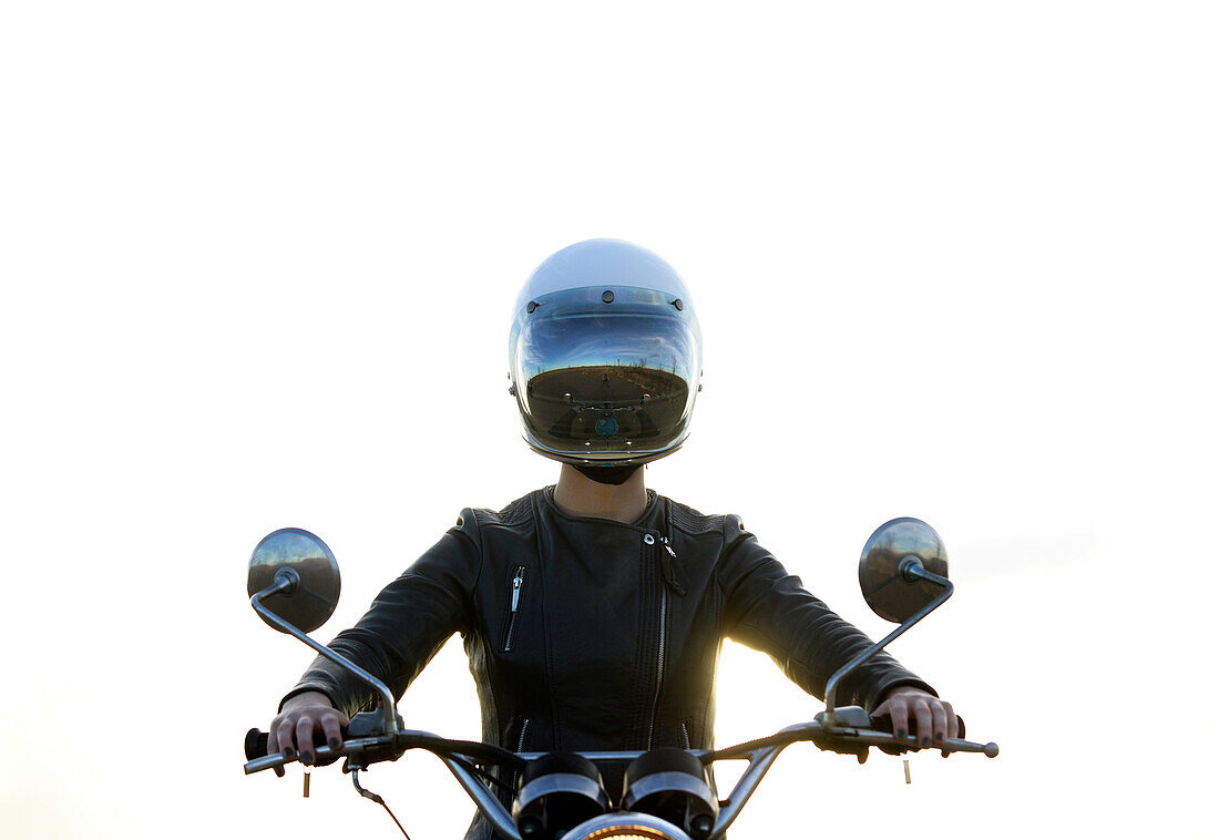 Centered perspective of a woman sitting on her vintage motorcycle wearing a reflective helmut face shield with a clean white background