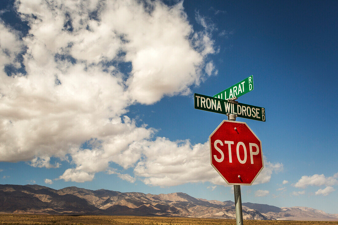 A stop sign and two street signs in the bottom corner of image, with desert mountains on horizon and a big, blue sky with puffy clouds