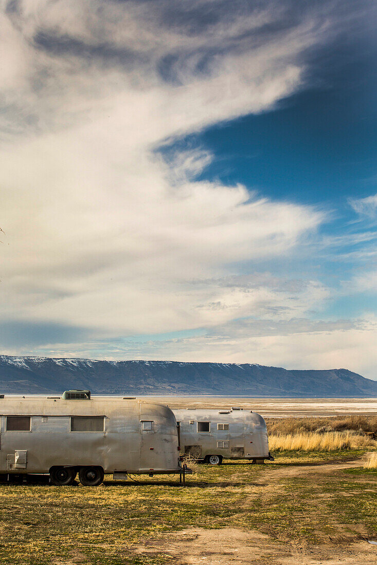 Two vintage Airstream trailers parked in a vast, dry landscape with blue sky and clouds above