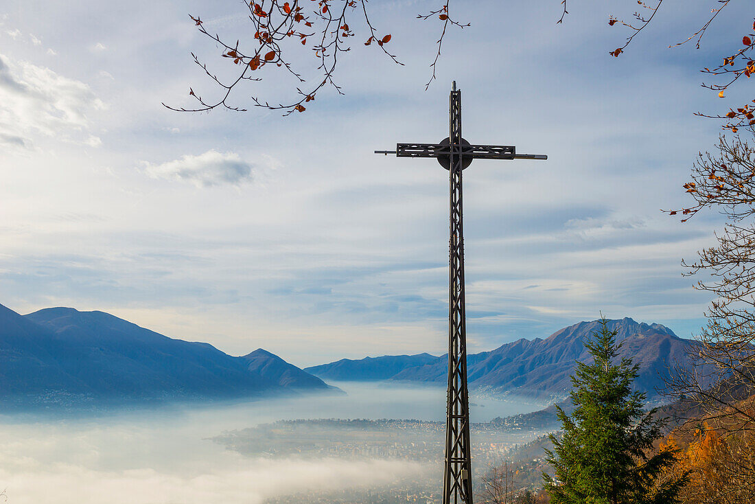 'A cross in the foreground and fog over a city and alpine lake; Locarno, Ticino, Switzerland'