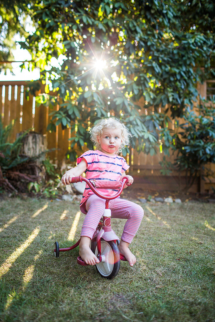 A blond toddler girl in pink sits on a tricycle in a backyard with sunlight streaming through the trees.