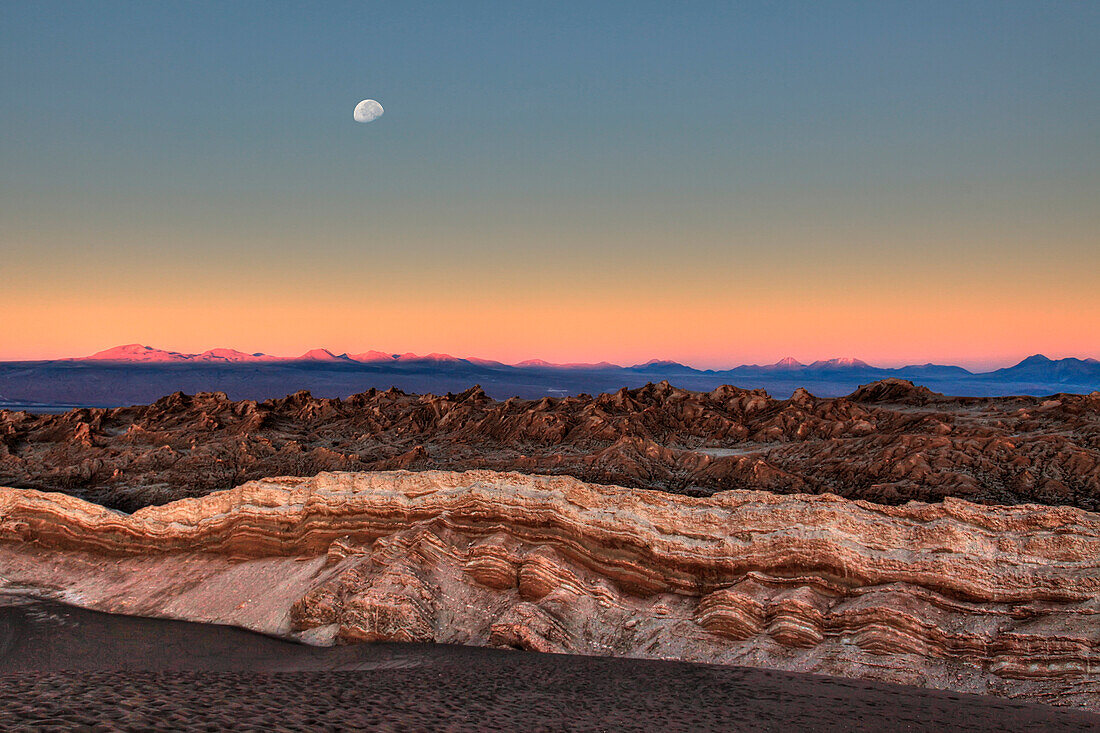 The moon appears above the peaks that surround the Valley of the Moon, illuminated by the light of sunset. Atacama Desert. Chile. South America