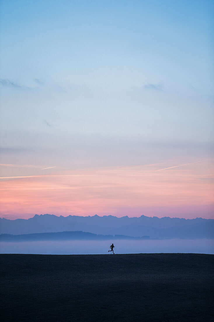 Junger Mann läuft über eine Wiese bei Sonnenaufgang, Allgäu, Bayern, Deutschland