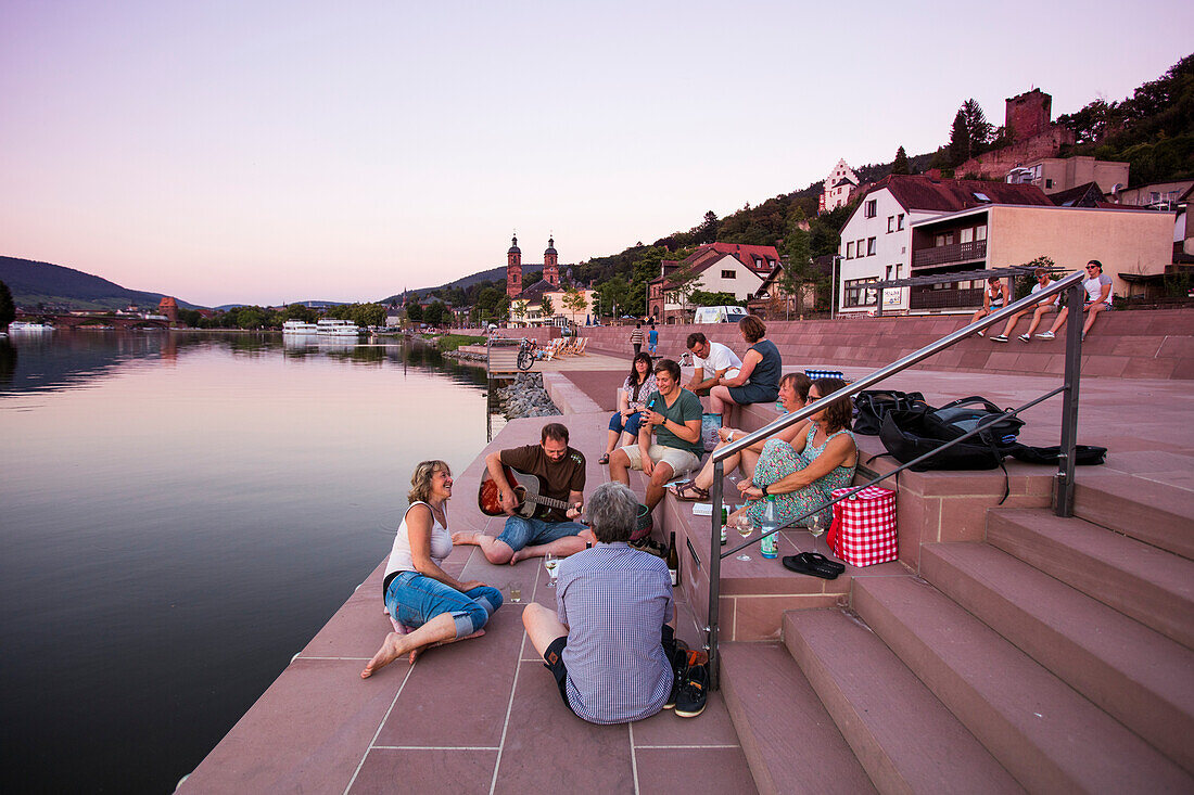 People sing, drink and relax on steps along riverfront of Main river at sunset, Miltenberg, Spessart-Mainland, Bavaria, Germany