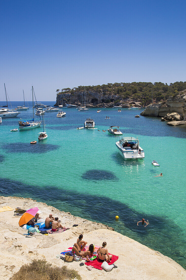 Menschen relaxen auf Felsen an der Bucht Cala Portals Vells mit vor Anker liegende Yachten und Segelboote, Portals Vells, Mallorca, Balearen, Spanien