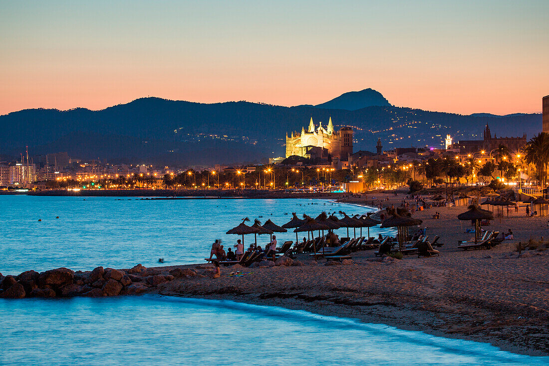 Silhouettes of people and thatched umbrellas at Nassau Beach Club with illuminated La Seu Palma Cathedral in distance at dusk, Palma, Mallorca, Balearic Islands, Spain