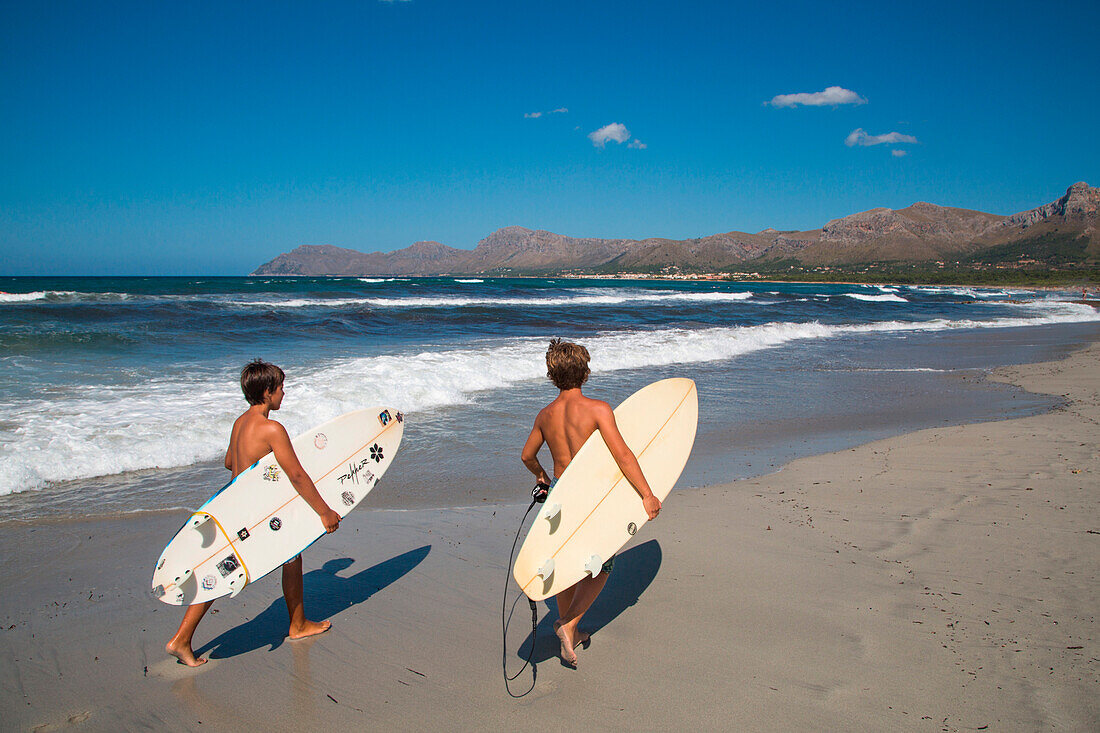 Zwei Jungen tragen Surfbretter am Strand der Bucht von Alcudia, Colonia de Son Serra de Marina, Mallorca, Balearen, Spanien