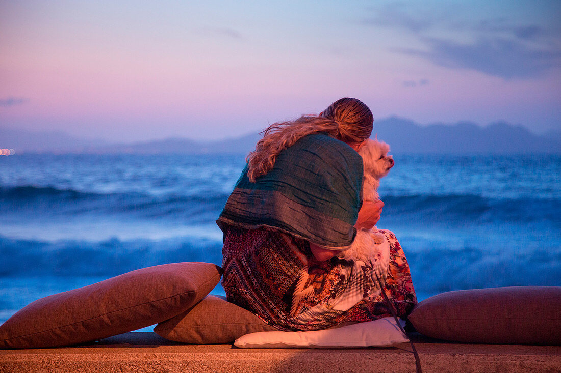 Young woman cuddles dog while sitting on cushions of Es Vivers restaurant on beach promenade with view of the Mediterranean Sea at dusk, Colonia de Sant Pere, Mallorca, Balearic Islands, Spain