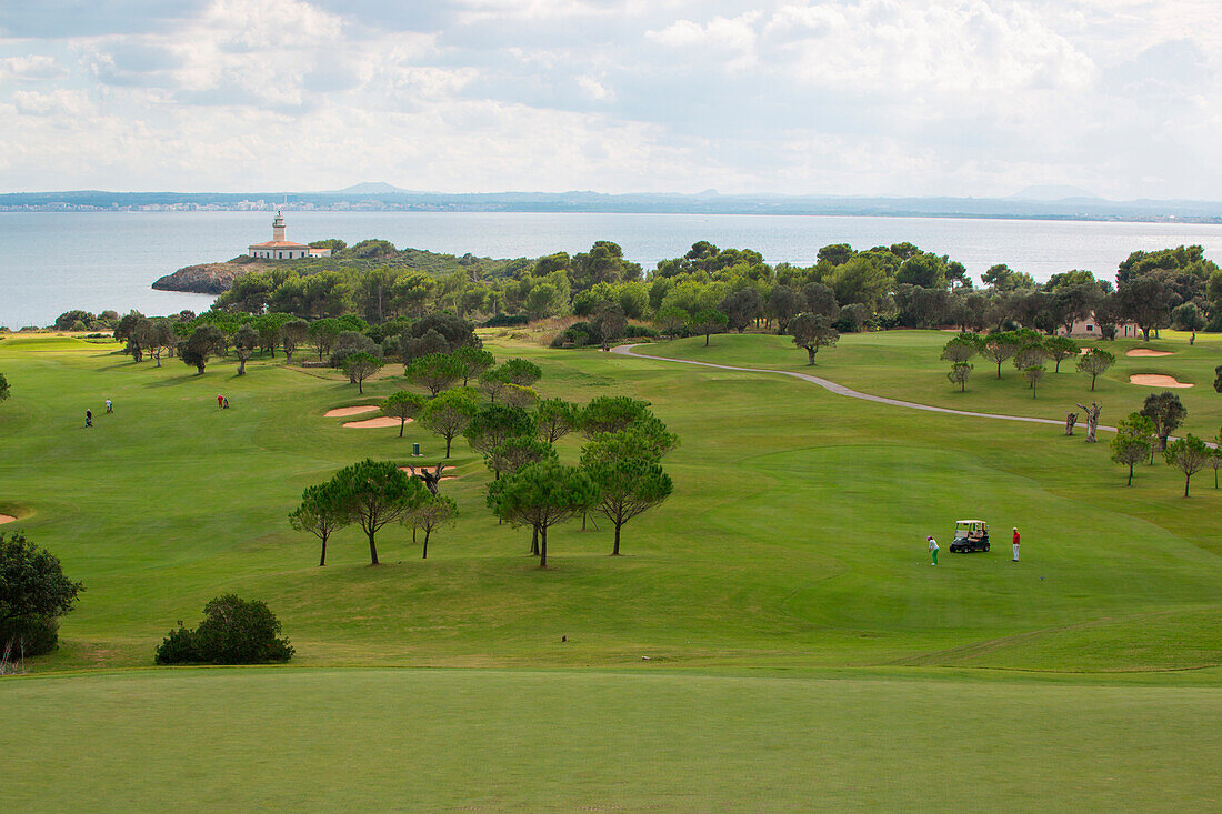 Golfers approach hole 12 at Club de Golf Alcanada with Faro de Alcanada lighthouse in distance, near Port d'Alcudia, Mallorca, Balearic Islands, Spain