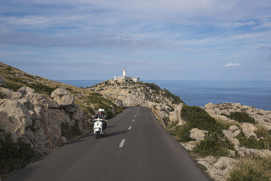 Paar auf weißer Vespa auf Straße entlang der Halbinsel Cap de Formentor mit Leuchtturm Faro de Formentor im Hintergrund, Cap de Formentor, Mallorca, Balearen, Spanien