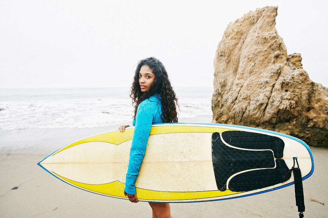Serious Mixed Race woman carrying surfboard at beach