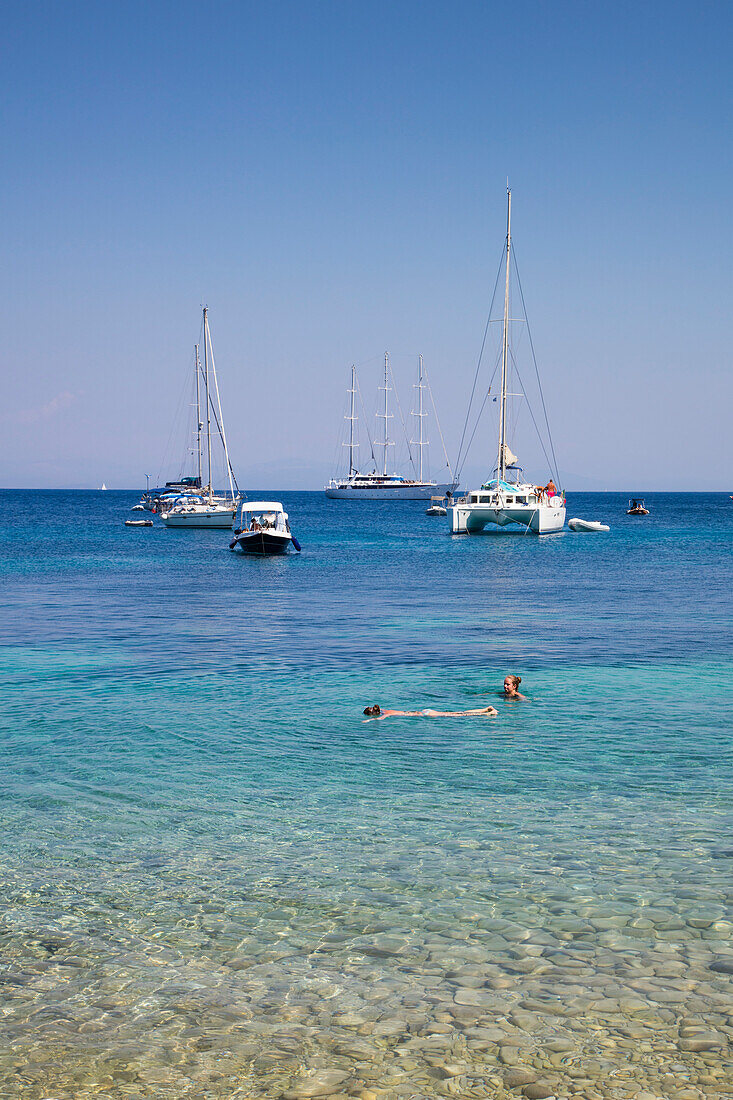 People swim in clear waters along pristine bay with sailboats and motor sailing cruise ship M/S Panorama (Variety Cruises) at anchor in distance, Paxos, Ionian Islands, Greece