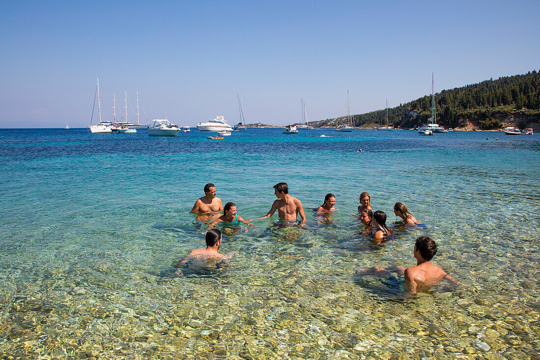 Gruppe von Menschen beim planschen und relaxen im Wasser nahe Strand mit Segelbooten im Hintergrund, Paxos, Ionische Inseln, Griechenland