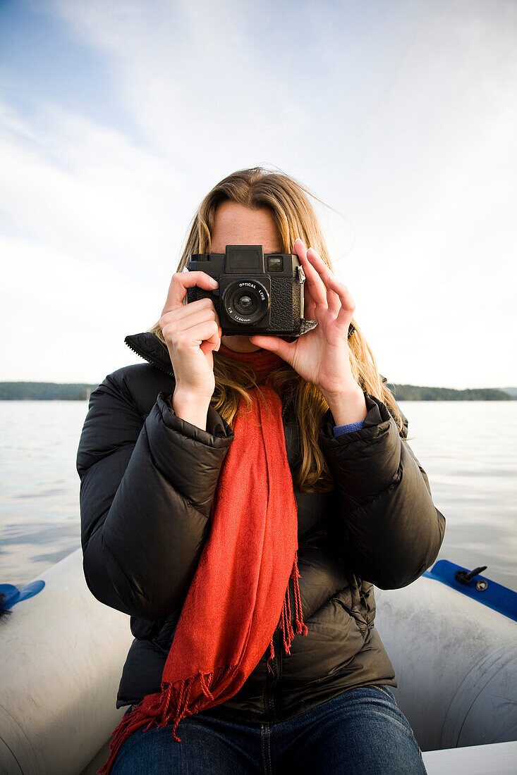 Young woman in red scarf and black jacket poses with vintage camera in an inflatable dingy afloat at sunset.