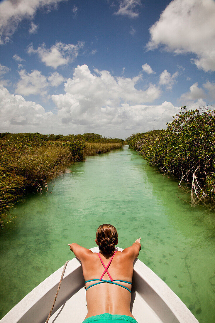 A young woman in a bikini sits in the bow of a white boat as it moves through teal-green water of the Sian Ka'an Preserve.
