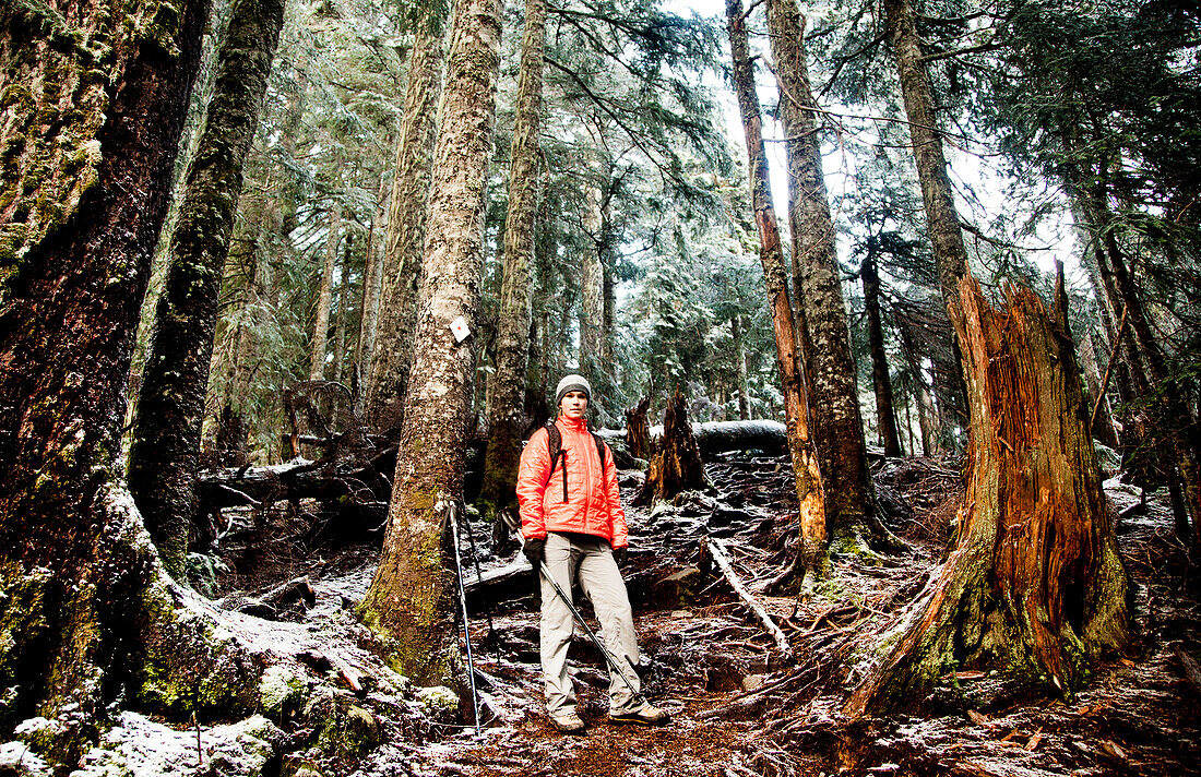A young woman hiker in a bright orange jacket pauses on a steep trail surrounded by tall fir trees on a cold winter day.