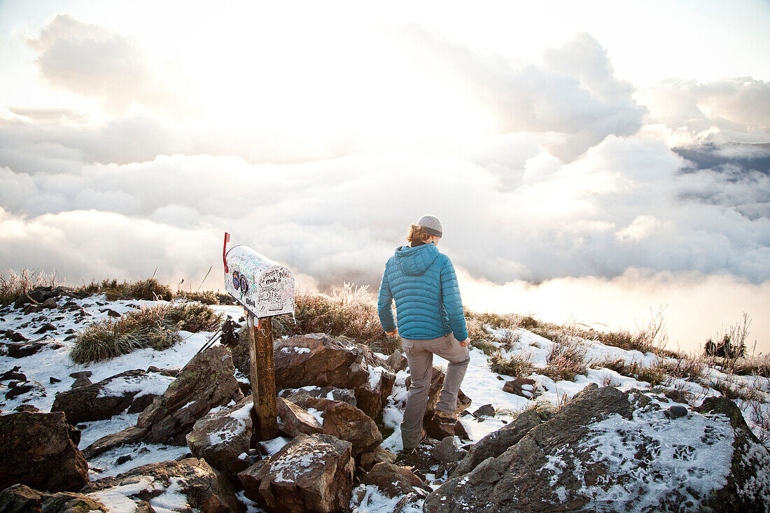 A young woman in a teal down jacket hikes among snowy rocks and near a mailbox at the top of Mailbox Peak in the Cascade foothills.