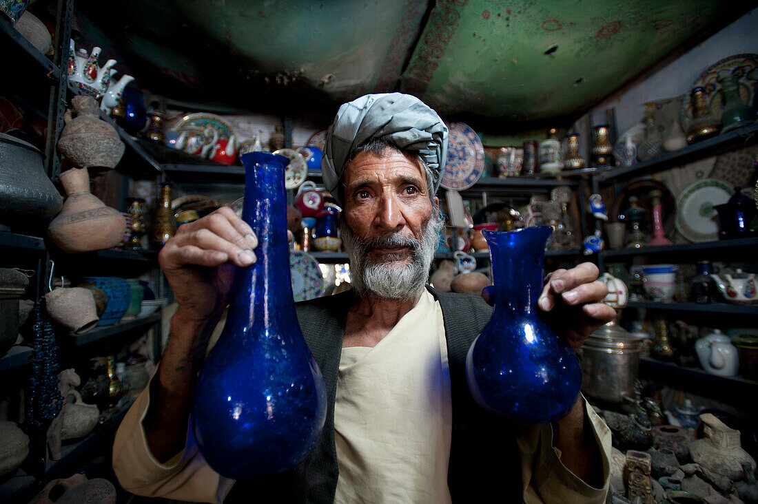 A glass blower holds up blue glass gourd-shaped vases in a trinket shop in Herat, Afghanistan, Asia