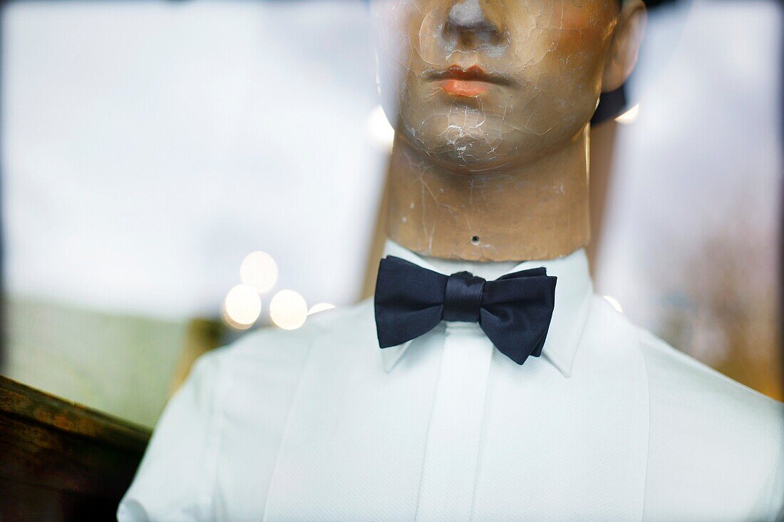 Portrait of an old mannequin seen through a shop window with reflections in the glass, smartly dressed in shirt, hat and bow tie
