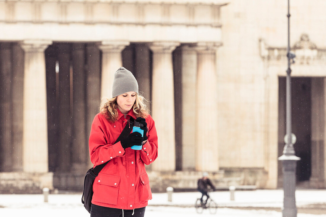 Junge Frau mit Isolierbecher am verschneiten Königsplatz in München, Bayern, Deutschland