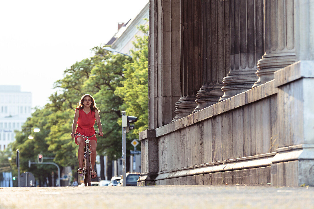 Young woman bicycling through the propylaea, Königs Plaza in Munich, Bavaria, Germany