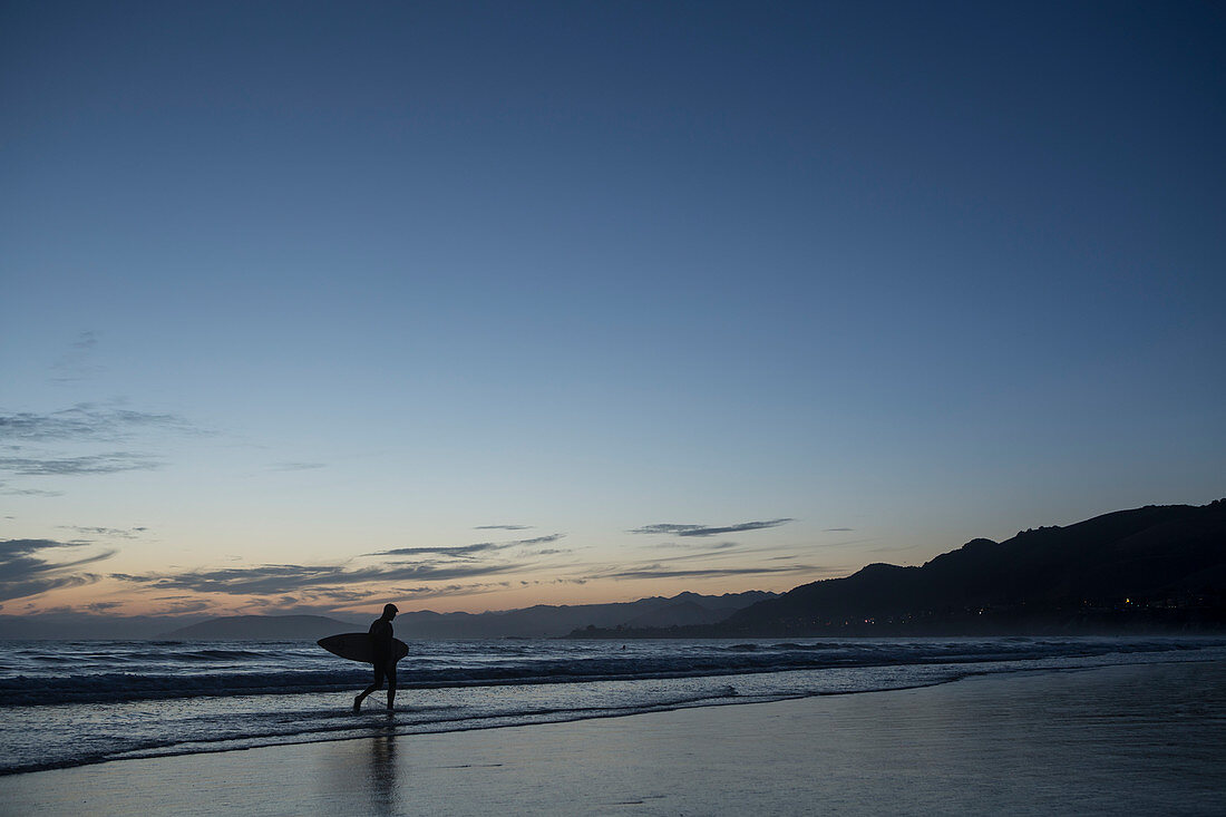 Man walking on beach with surf board, San Luis Obispo, California, United States of America
