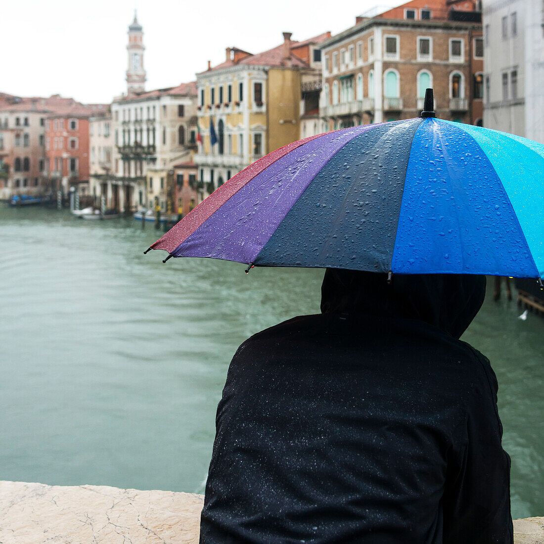 A person standing with a wet, colourful, umbrella looking out over a canal, Venice, Italy