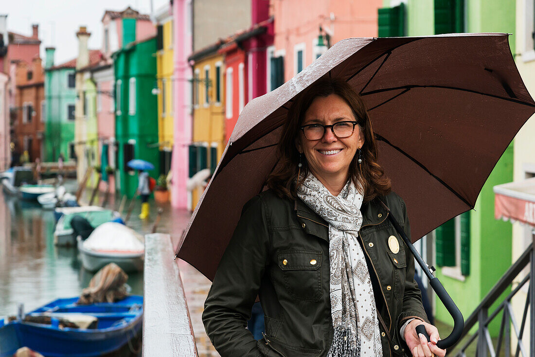 A woman stands beside a canal with an umbrella with colourful houses in a row in the background, Venice, Italy