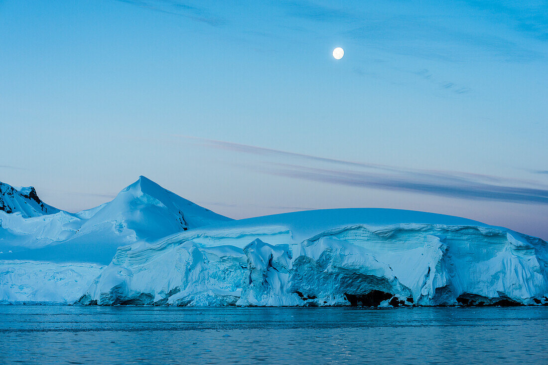 Full moon in a blue sky over the snow covered mountains and blue ocean, Antarctica