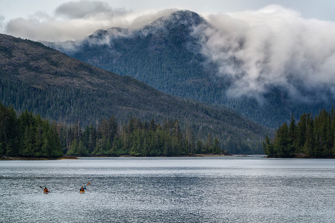 Two kayakers paddling toward the mainland while clouds flow over the coastal mountains, Haida Gwaii, British Columbia, Canada