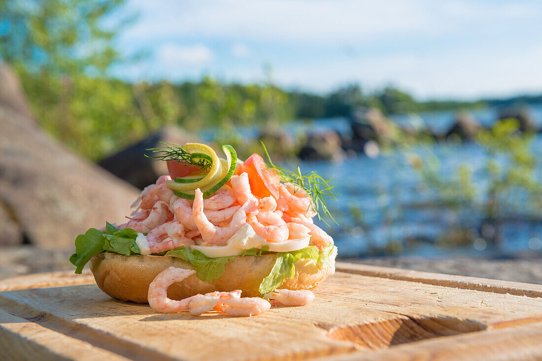 Bun with shrimps, egg, cucumber and lemon on a wooden plate in front of lake Vanern, Smaland, Sweden