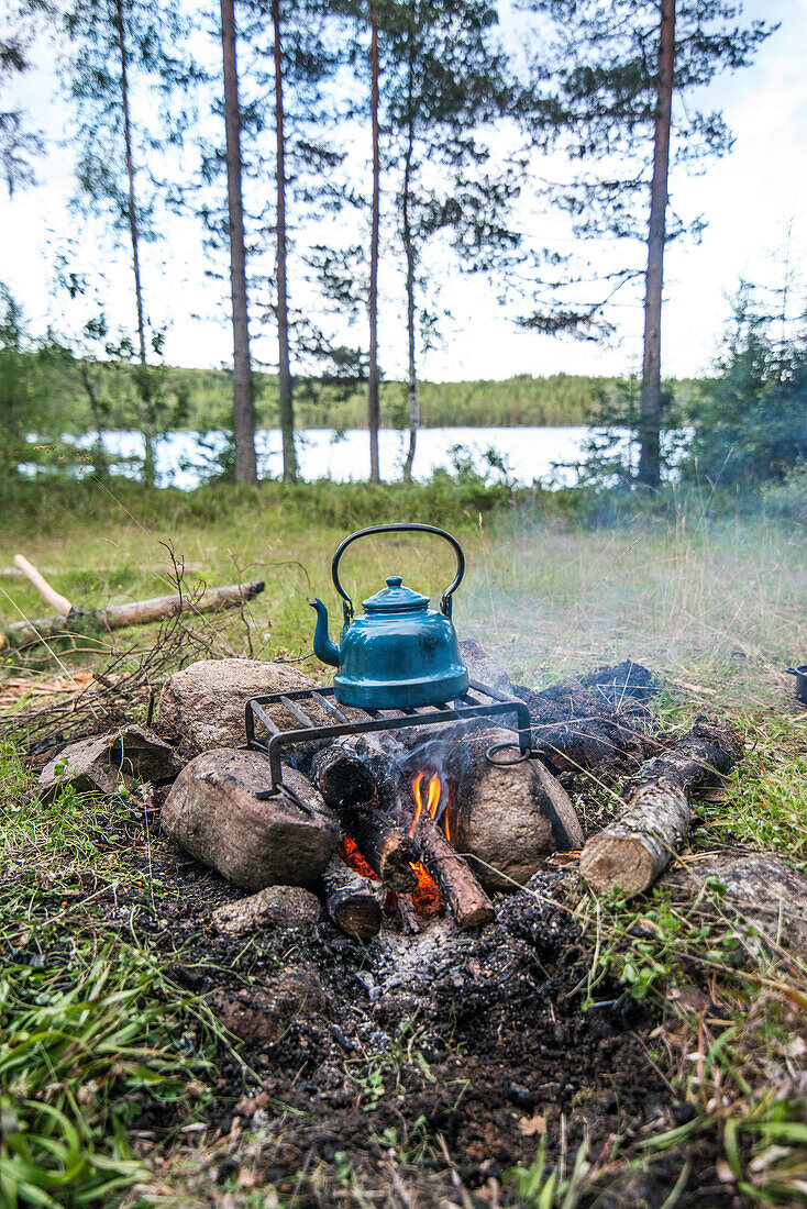 Blauer Wasserkessel aus Emaille steht auf einem Rost über dem Lagerfeuer am See, Värmland, Schweden