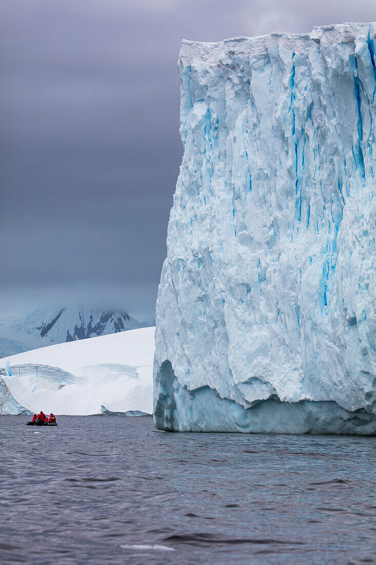 Ein Zodiac Schlauchboot von Expeditions Kreuzfahrtschiff MV Sea Spirit (Poseidon Expeditions) im Größenkontrast zu einem riesigen Eisberg, Guyou Inseln, Flandres Bay, Grahamland, Antarktische Halbinsel, Antarktis