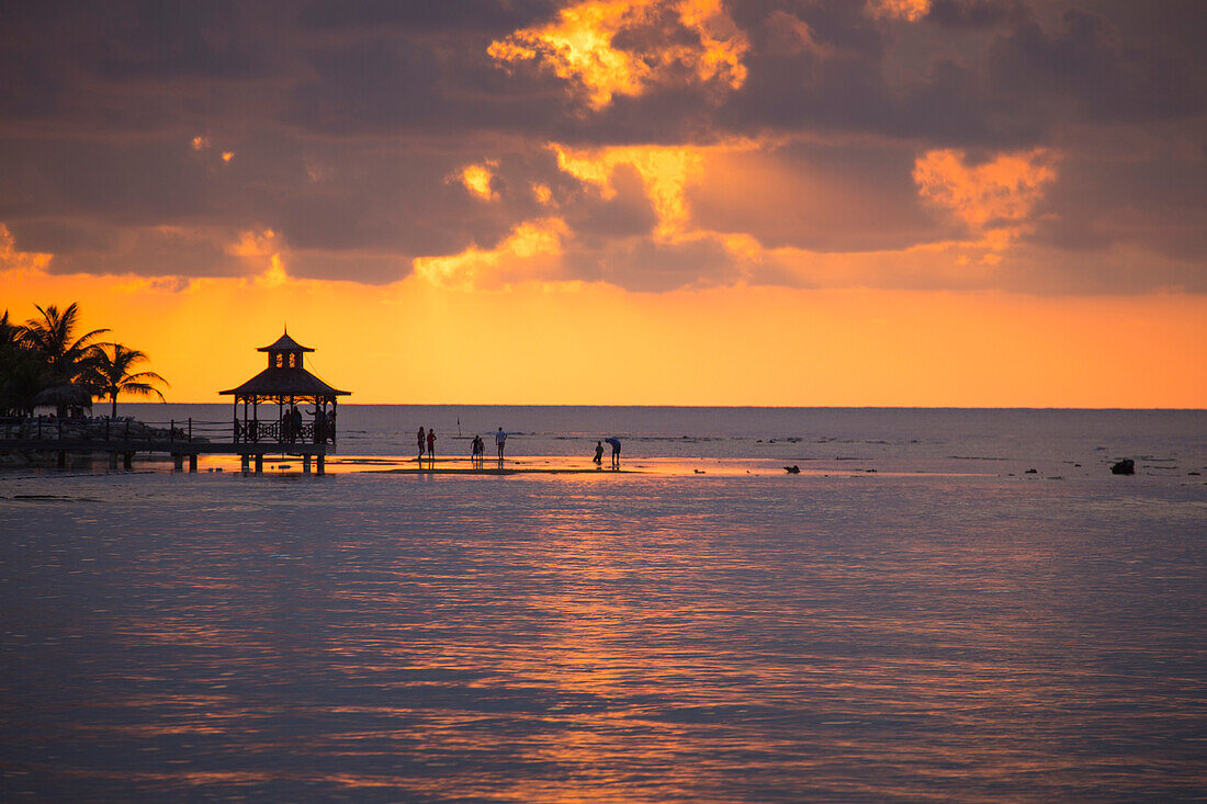 Silhouette von Menschen im flachen Wasser und Pavillon auf Steg bei Sonnenuntergang, Rose Hall, nahe Montego Bay, Saint James, Jamaika