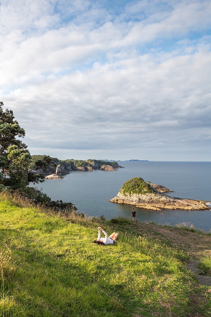 Hahei, Mercury Bay, Küstenlandschaft, Frau liegt im Gras, smartphone, Coromandel Peninsula, Nordinsel, Neuseeland
