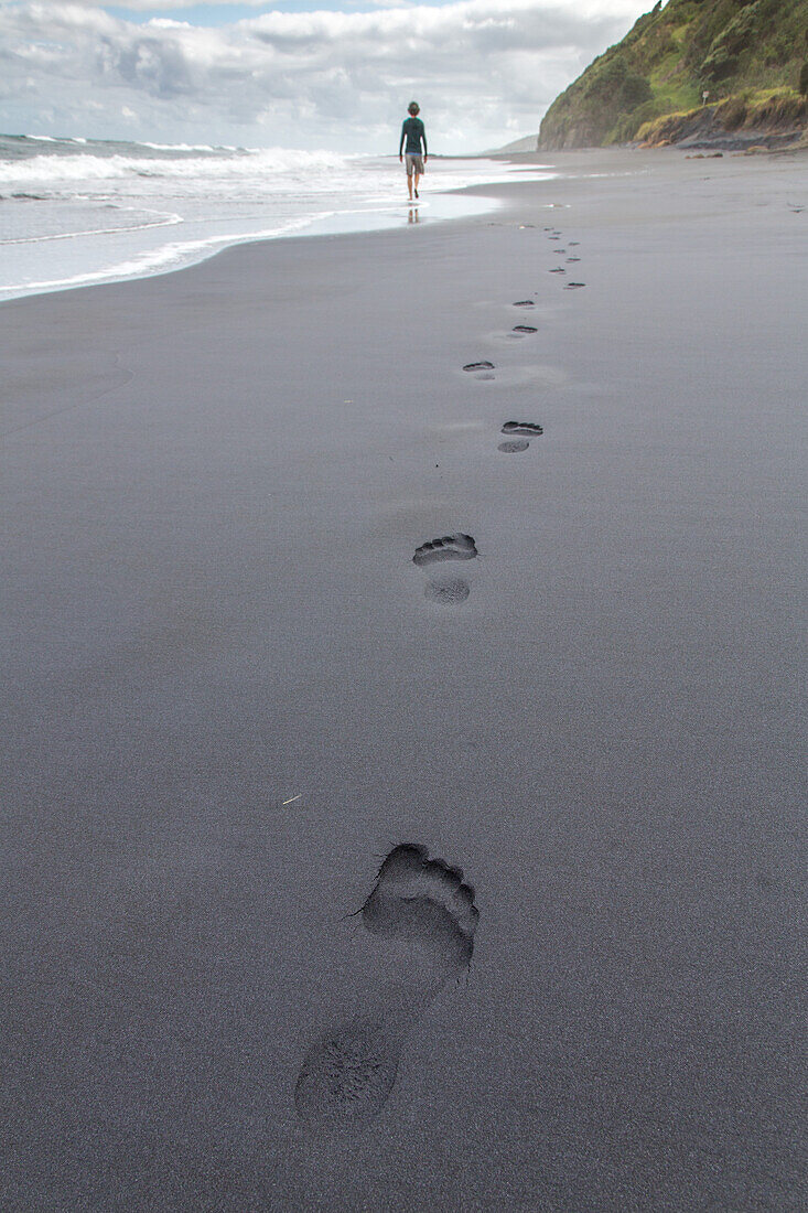 footprints in black volcanic sand on beach with person, barefoot, high format, North Island, New Zealand