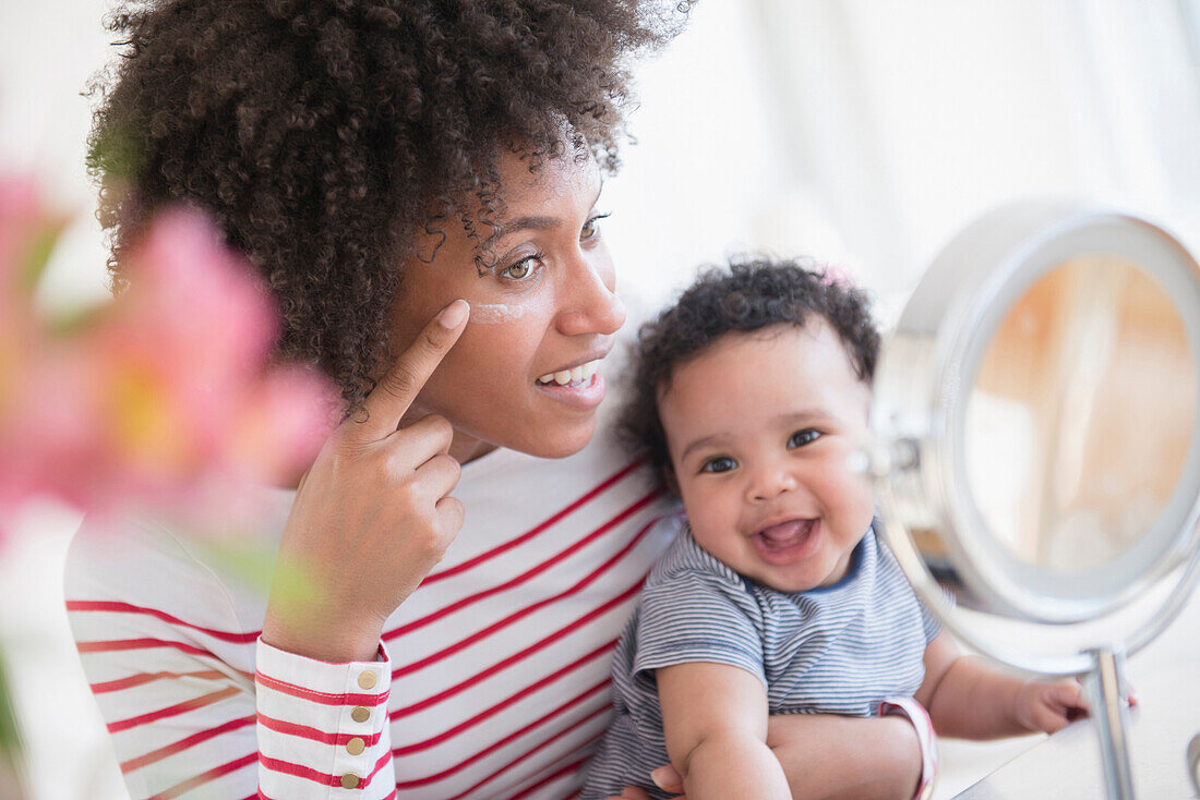 Mother holding baby son applying face cream in mirror