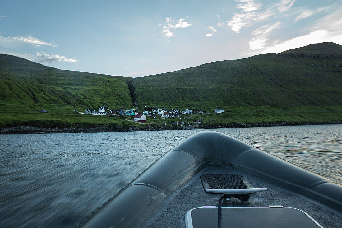 Blick von einem Boot auf ein kleines Dorf an einem grün bewachsenen Berg, Färöer Inseln