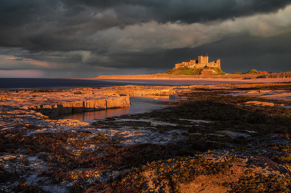 A storm passes behind Bamburgh Castle with the last light of the day illuminating the rocky shoreline and castle, Northumberland, England, United Kingdom, Europe