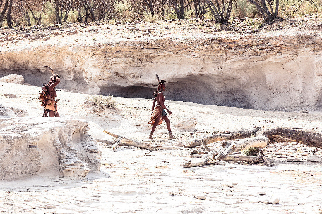 Zwei Himbafrauen, eine davon mit ihrem Kind am Rücken, suchen ihre Rinder in einem Trockenfluss, Kunene, Namibia