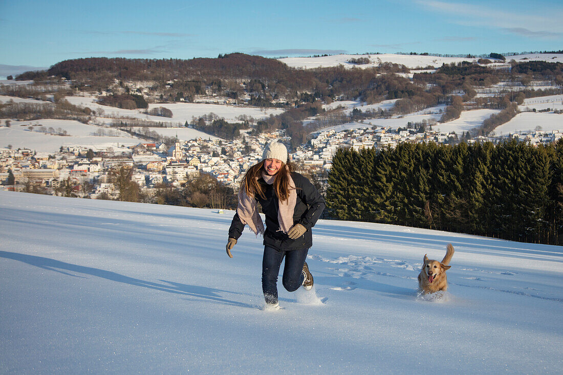 Happy teenage girl and golden retriever dog run through snow with view of Hilders in winter, Hilders, Rhoen, Hesse, Germany