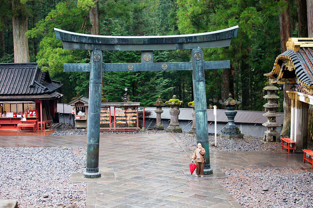 Alter Mönch steht mit rotem Regenschirm am Torii des Toshogu-Schrein, Nikko, Tochigi Präfektur, Japan