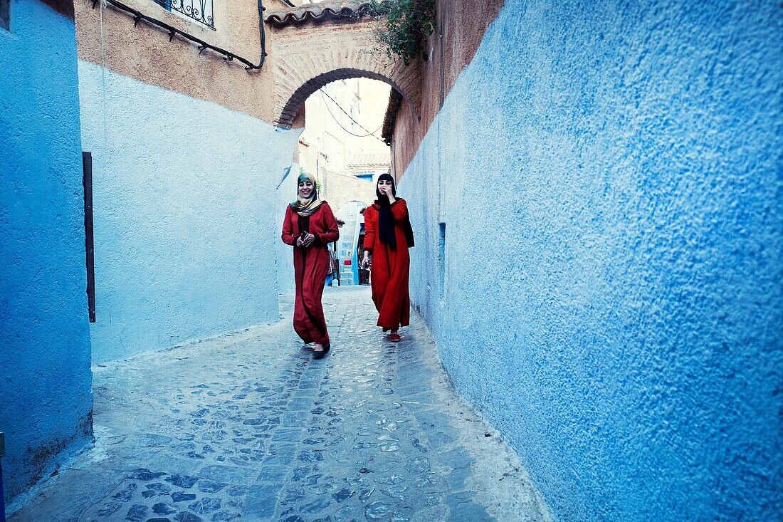 Two smiling Moroccan women walking in the medina, wearing red dresses. Chaouen, Morocco