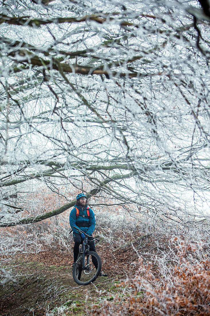 Young man standing with his bike in a with frost covered forest, Allgaeu, Bavaria, Germany