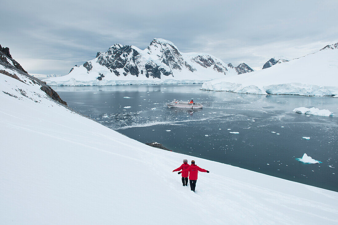 Zwei Menschen auf einem schneebedeckten Hügel mit Expeditions-Kreuzfahrtschiff MS Bremen (Hapag-Lloyd Cruises) im Hintergrund, Orne Harbor, Grahamland, Antarktische Halbinsel, Antarktis